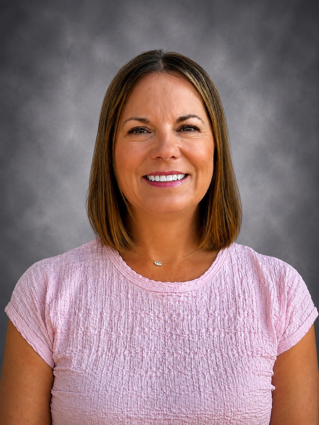 A woman with brown hair pulled back smiles at the camera, wearing a pink shirt.